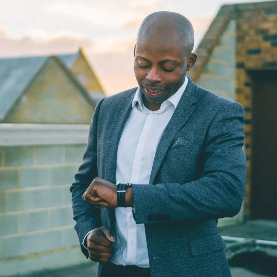 Man in a suit checking his watch against a rooftop backdrop at sunset.