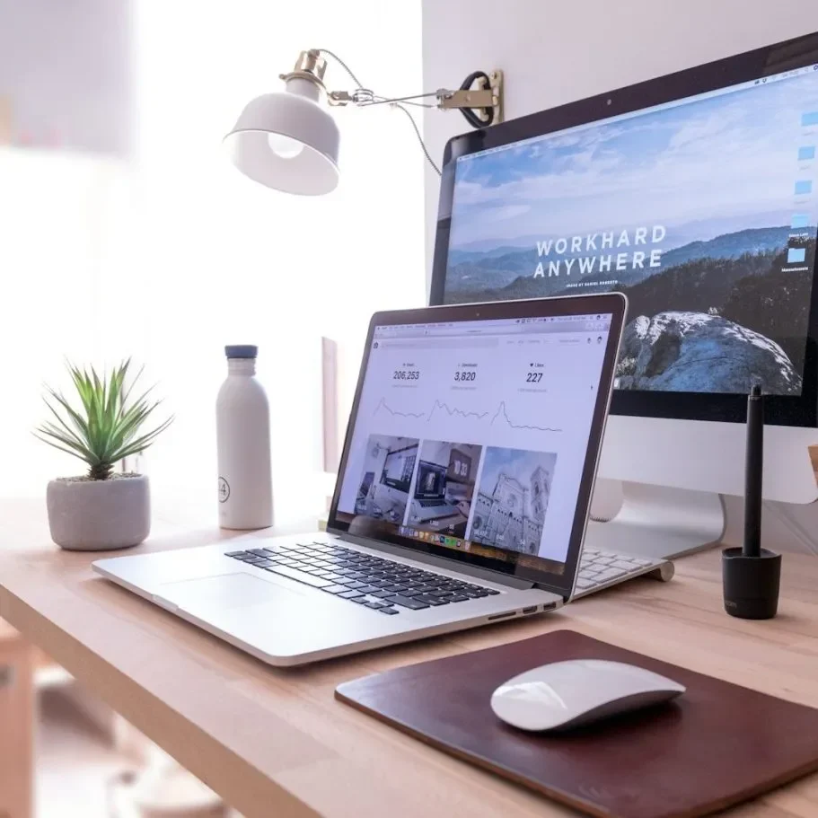 Modern workspace featuring a laptop, desktop monitor, plants, and a desk lamp.