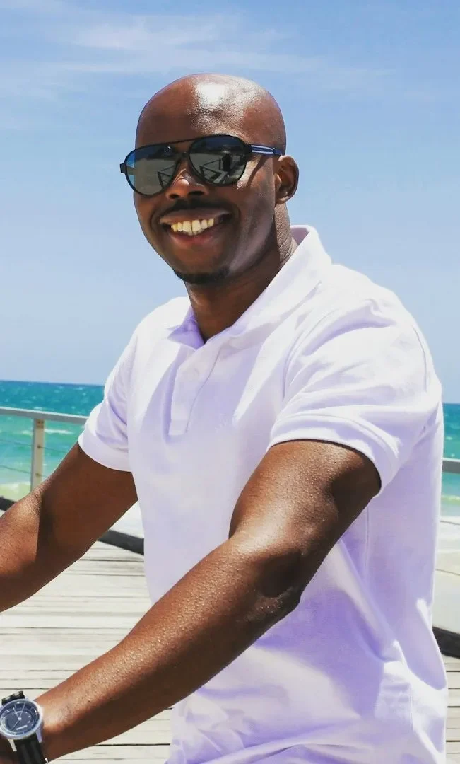Smiling man in sunglasses riding a bike along a beachside boardwalk.